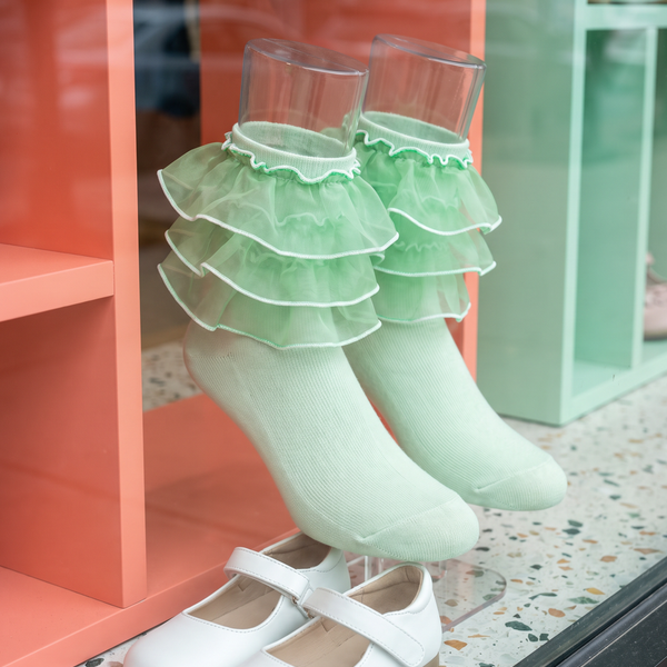 Pale-green girls' socks with four distinct organza ruffle tiers displayed beside white Mary Jane shoes.