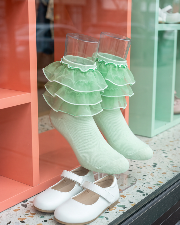Pale-green girls' socks with four distinct organza ruffle tiers displayed beside white Mary Jane shoes.