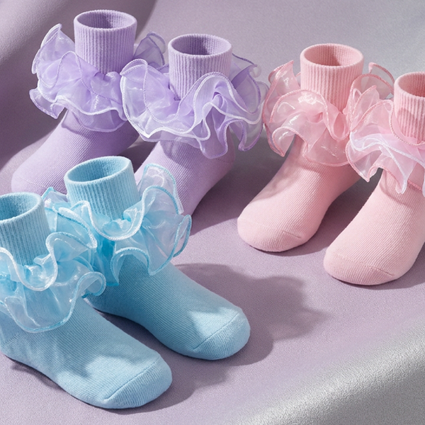 Three pairs of girls' layered ruffle socks in sky blue, lavender and pink arranged on satin fabric.