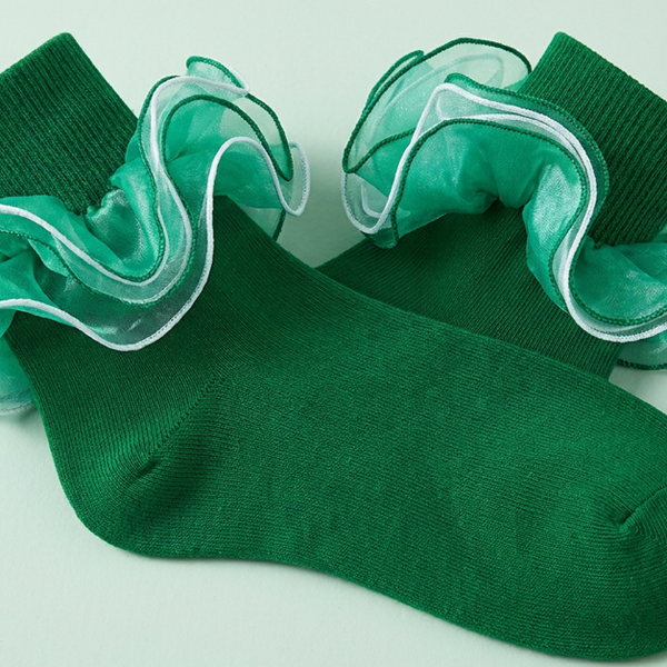 A pair of emerald green ankle socks with layered sheer ruffles shown close up on a pale mint surface.