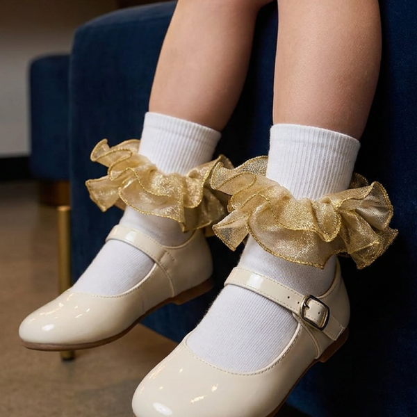A girl's white socks with shimmering gold ruffle trim paired with cream Mary Jane shoes in a dressing room.