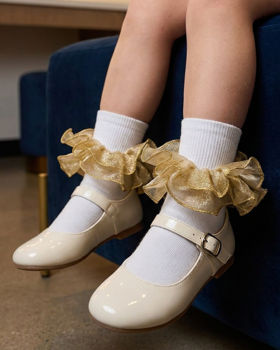 A girl's white socks with shimmering gold ruffle trim paired with cream Mary Jane shoes in a dressing room.