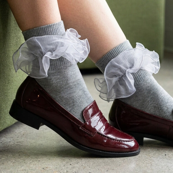 Grey socks with layered white ruffle cuffs styled with burgundy leather loafers beside a green sofa.