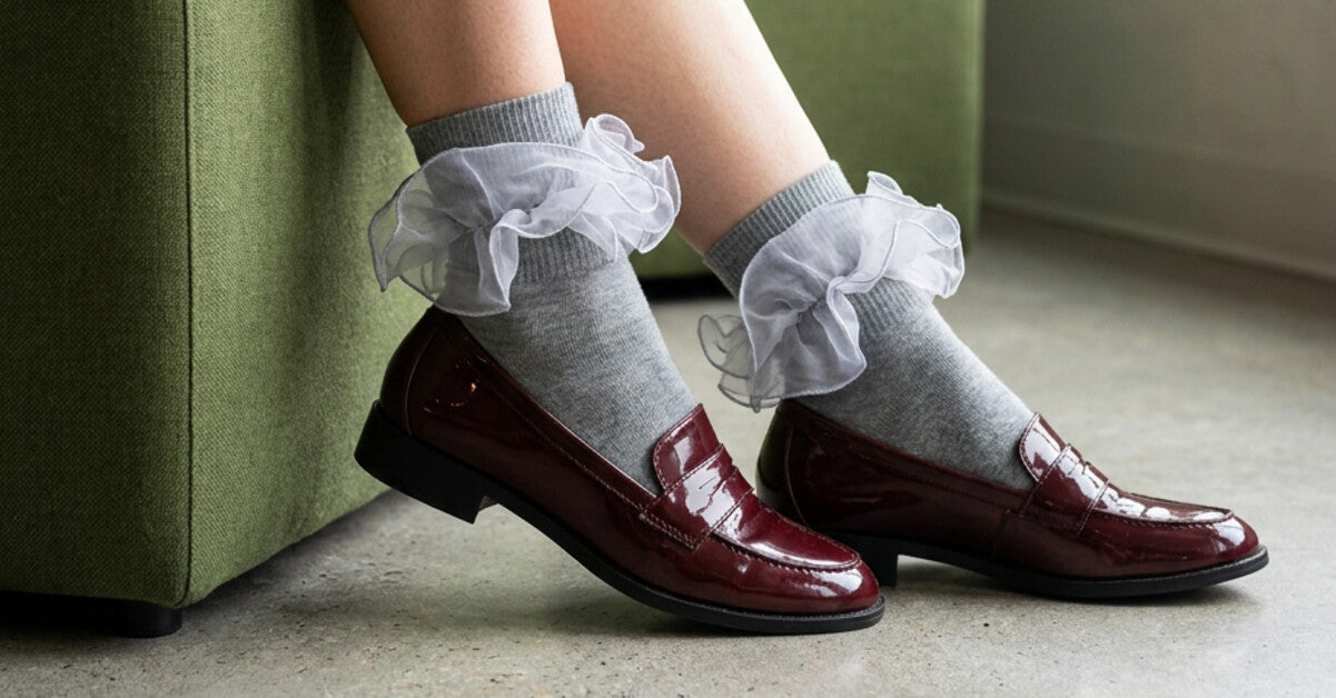 Grey socks with layered white ruffle cuffs styled with burgundy leather loafers beside a green sofa.